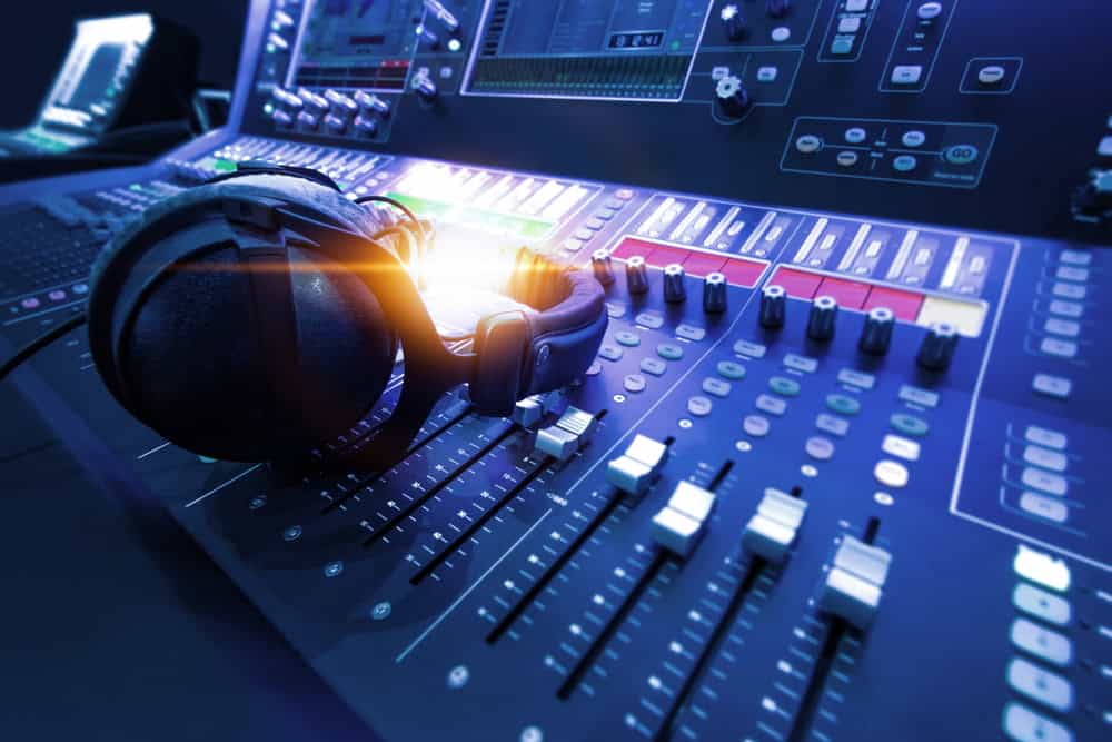 A pair of headphones rests on a professional audio mixing console. The board is sleek and illuminated, showcasing multiple faders, knobs, and digital screens. This high-tech ambiance in the dimly lit studio reminds us of the importance of E-waste disposal to sustain such futuristic environments.