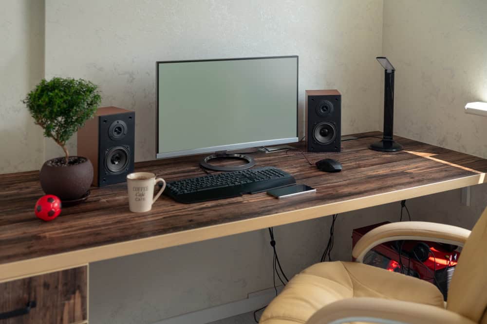 A tidy home office desk with a flat-screen monitor, keyboard, mouse, and speakers sits ready for productivity. A small potted plant made from recycled computers, coffee mug, and red ball adorn the wooden surface. A beige chair is nearby, with a floor lamp to the right.