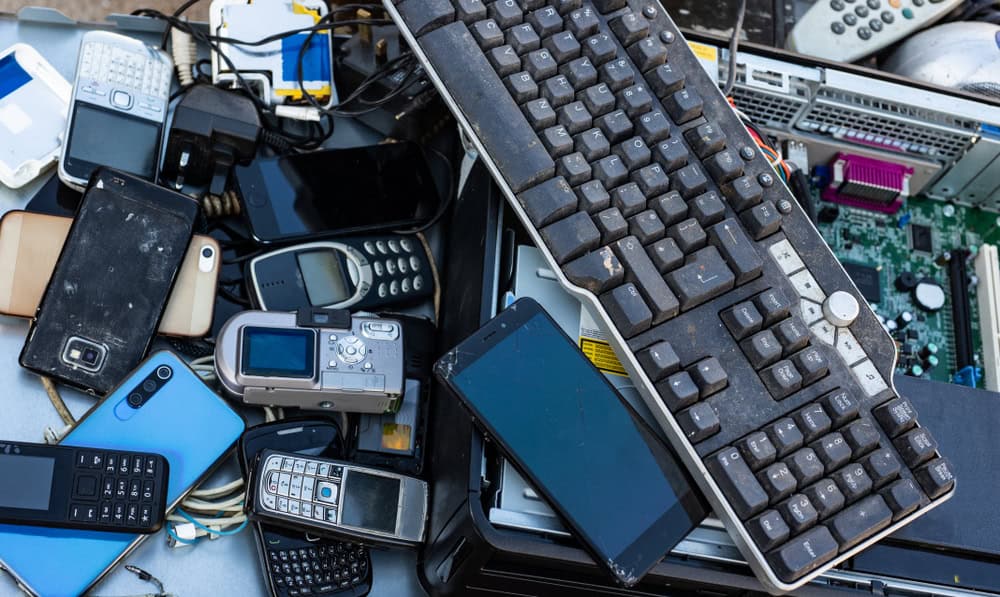 A pile of obsolete electronics, including various mobile phones, an old keyboard, a digital camera, and computer parts. These worn devices symbolize the growing issue of electronics waste disposal, highlighting the urgent need for responsible e-waste disposal solutions.