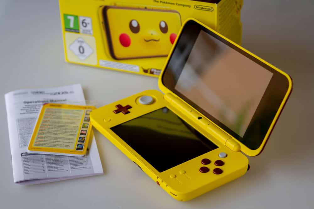 A yellow Nintendo 2DS XL with a Pikachu design is open on the table. Beside it, an instruction booklet and a card with game details sit ready. In the background, a matching yellow Pikachu-themed box hints at joy and sustainability&mdash;perfect for fans who recycle electronics responsibly.