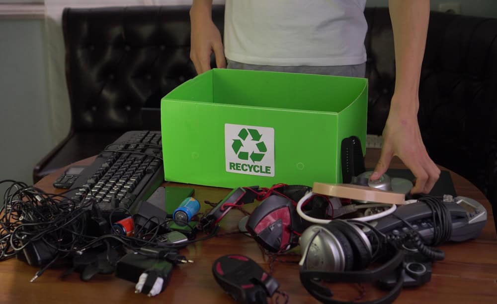 A person is organizing electronics waste for recycling, neatly placing a keyboard, headphones, chargers, and various cables into a green box adorned with the recycling symbol. The setup is on a table with a black couch in the background, highlighting their commitment to responsible electronics disposal.