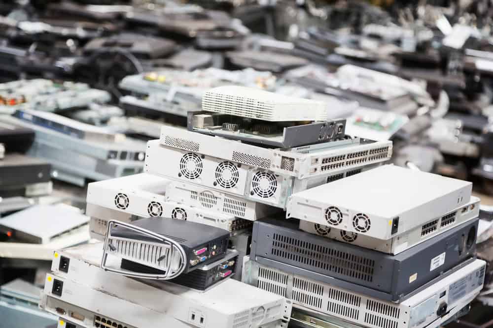 A large pile of discarded electronic equipment, including servers and network devices, is stacked in an E-waste disposal facility. Among the e-waste are components like recycle laptops, visible in varying sizes and shapes with fan vents and ports on some units.