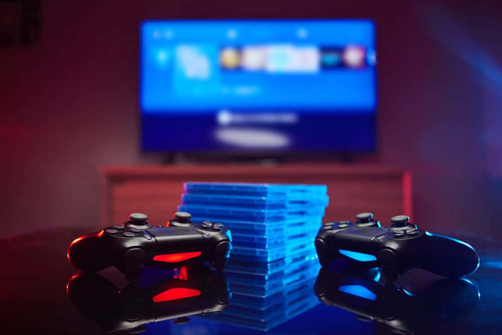 Two gaming controllers rest on a reflective surface, one glowing red and the other blue. Between them, a stack of game cases sits as a reminder of cherished adventures. The TV screen displays a vivid gaming interface, while dim lighting enhances the atmosphere&mdash;an invitation to recycle electronics thoughtfully.