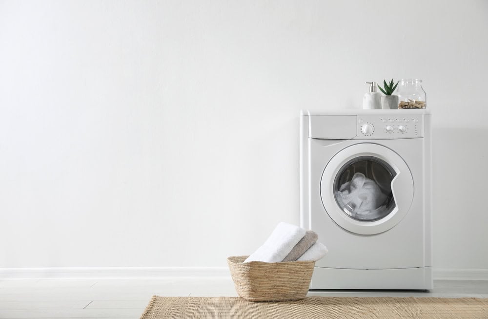 A white washing machine stands against a plain light-colored wall, embodying the essence of electronics recycling. A woven basket with folded towels sits on a beige rug in front, while a small plant and some jars on top complete the clean and minimalistic setting.