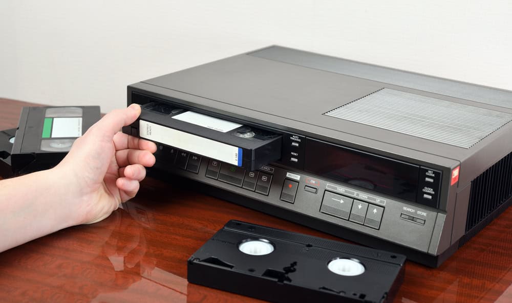 A person inserts a VHS cassette into a vintage VCR player on a wooden surface, embracing nostalgia before considering electronics recycling. Another VHS cassette lies next to the player, showcasing its spools. The VCR, with its array of control buttons, stands as a reminder of evolving technology and E-waste disposal challenges.