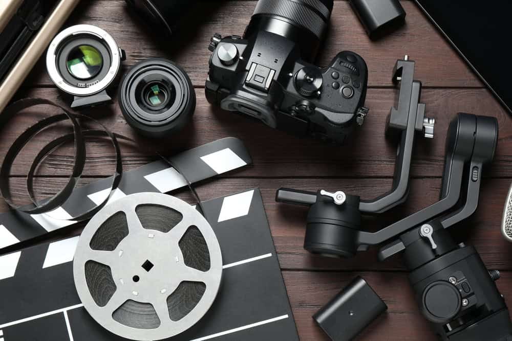 A flat lay of film and photography equipment on a wooden surface, including a camera, lenses, a film reel, a clapperboard, and a camera stabilizer. The thoughtfully arranged gear serves as a reminder to recycle electronics like old cameras and laptops to reduce waste in the filmmaking world.