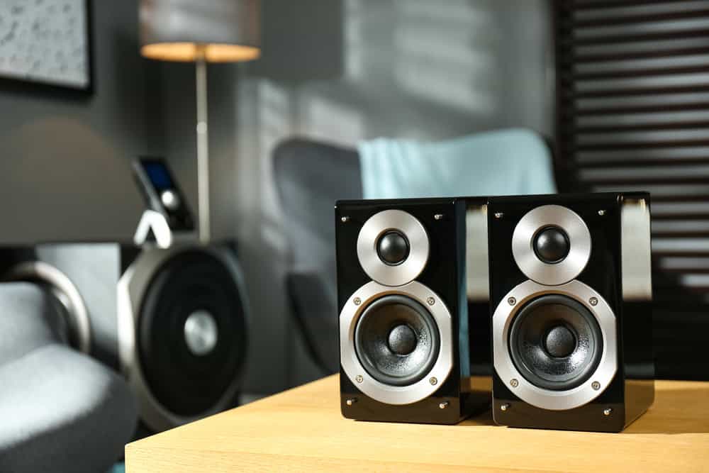 Two black and silver speakers sit on a wooden table in a modern living room. In the blurred background, a stylish floor lamp, a large black speaker, and a touch of blue fabric draped over a chair can be seen. Consider electronics recycling when upgrading your audio setup.