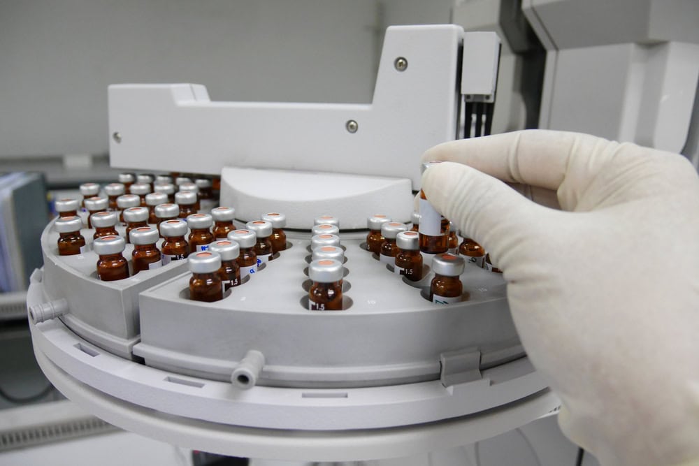 A gloved hand is placing a small vial of amber liquid onto a circular tray filled with similar vials in a laboratory dedicated to E-waste disposal. The tray is part of a white, automated machine used for handling samples crucial to electronics recycling research.