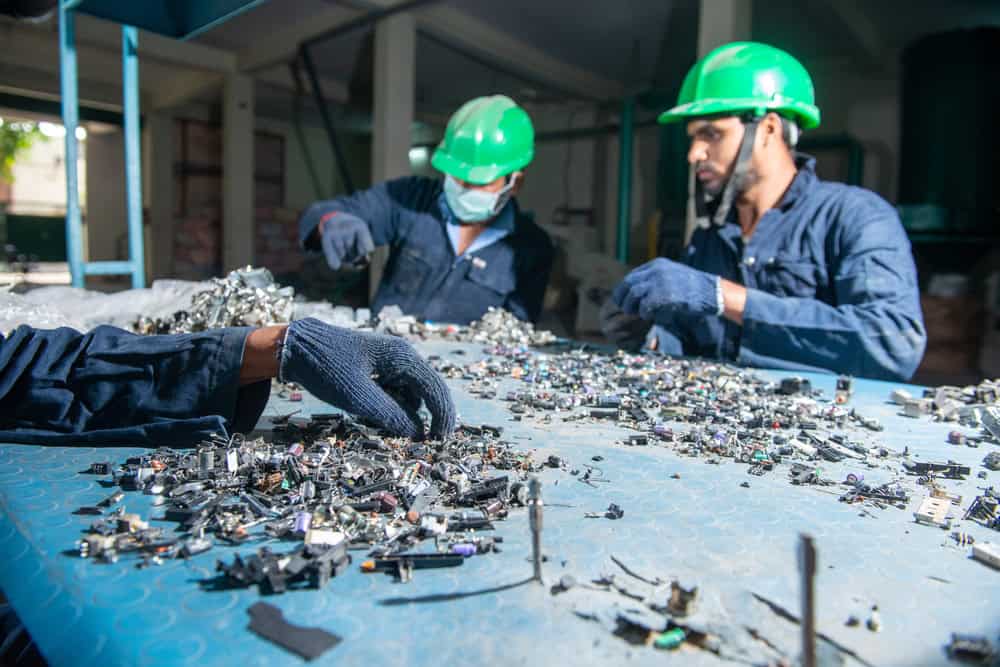 Workers wearing green helmets are diligently sorting through electronic waste on a blue table, exemplifying effective e-waste disposal. One worker reaches for small components while another focuses intently, emphasizing the meticulous process involved in electronics recycling against an industrial backdrop.