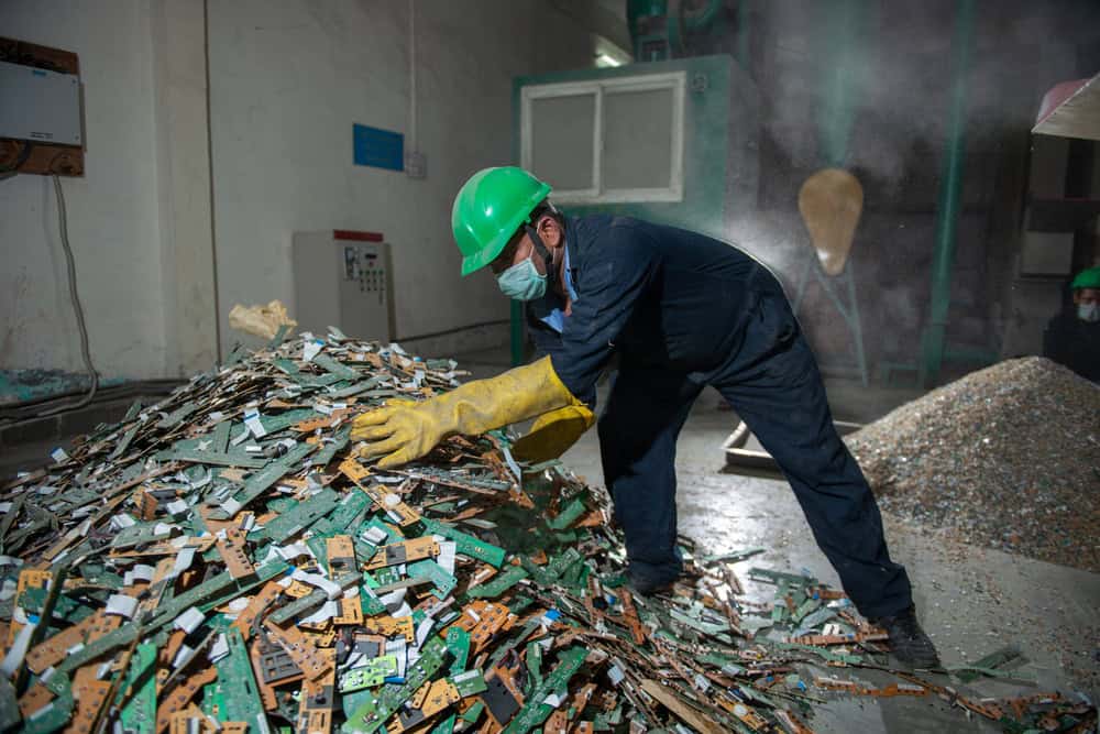 A worker wearing a green helmet, face mask, and yellow gloves navigates the dust-filled air to sort through a large pile of electronic waste for electronics recycling in an industrial setting. As he carefully examines circuit boards, another worker is visible in the background.