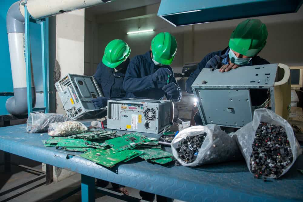 Three workers in green helmets and protective gear dismantle electronic equipment on a blue workbench. Bags of electronic waste and circuit boards are visible, indicating a recycle laptops or computer recycling process.