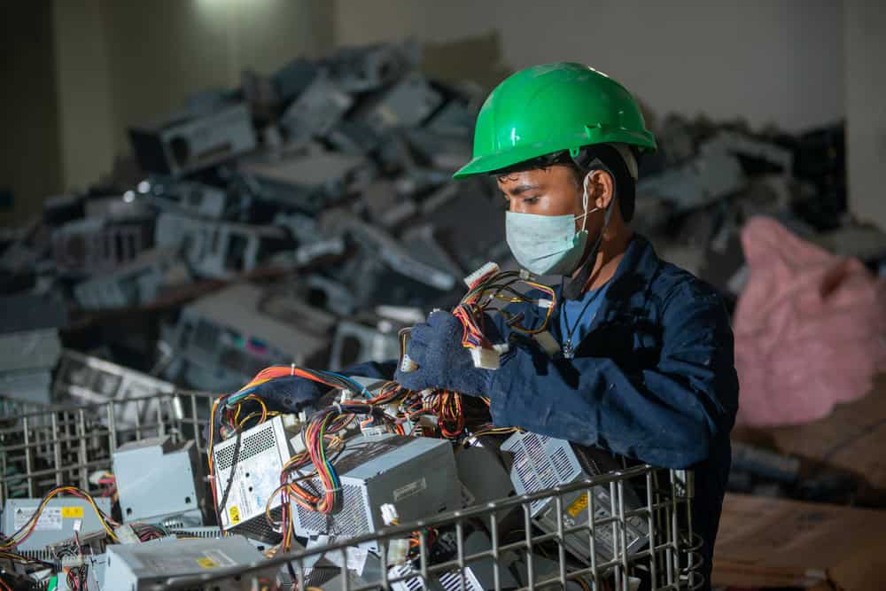 A worker wearing a green helmet and a mask inspects electronic waste, focusing on cables and components, in a dimly lit setting. Surrounded by discarded electronics, this scene highlights the crucial process of electronics recycling.