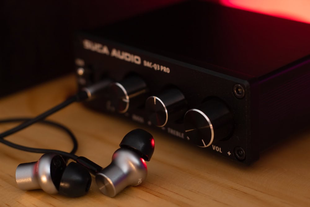 Close-up of a black audio amplifier with labeled knobs for bass, treble, and volume. A pair of silver and black earphones is plugged into the device, resting on a wooden surface. With emphasis on electronics waste disposal, the softly lit background shines in a warm red hue.
