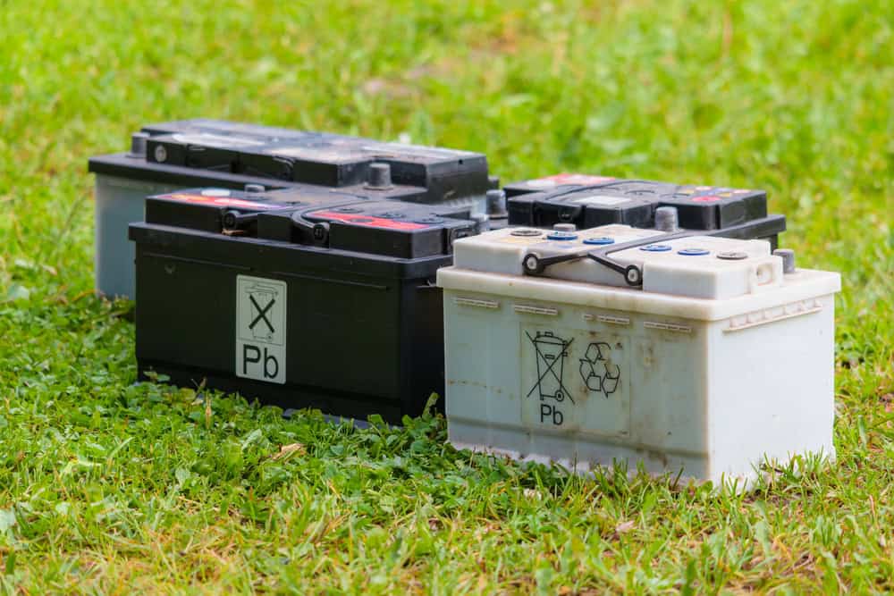 Four lead-acid car batteries, two black and two white, are placed on green grass. The batteries bear recycling and warning symbols, emphasizing the importance of electronics waste disposal due to their lead (Pb) content.