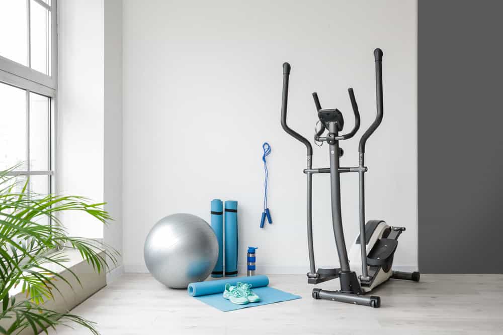 A home gym setup with an elliptical machine, two blue yoga mats, a silver exercise ball, a jump rope, and blue sneakers on a mat. A potted plant is partially visible near a window, where you're reminded to think of E-waste disposal when upgrading any electronics.