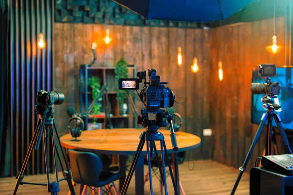 A cozy podcast studio with a round wooden table, surrounded by microphones and three video cameras on tripods. Warm lighting illuminates the room, while wall shelves with plants create a relaxed ambiance&mdash;perfect for discussions on electronics recycling and sustainable practices.