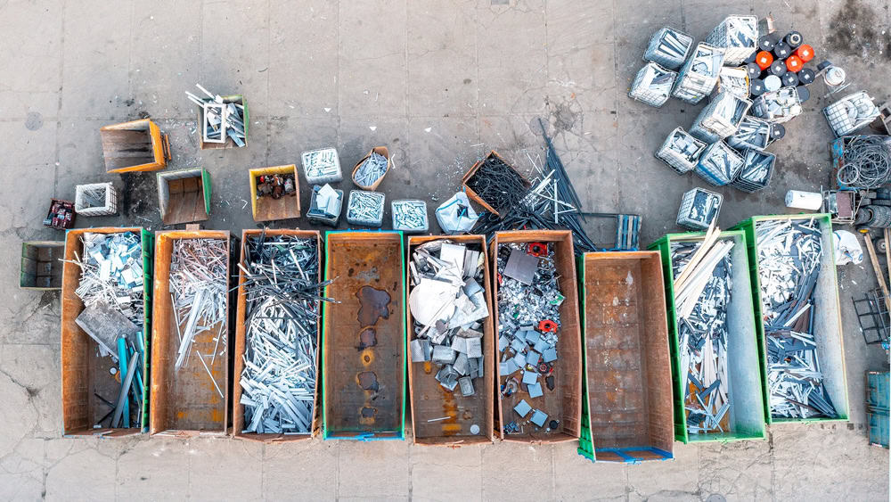 Aerial view of multiple large open-top containers filled with various types of metal scraps, ideal for electronics waste disposal. The scene includes rods, sheets, mixed pieces, and smaller bins alongside metal coils scattered on a concrete surface.
