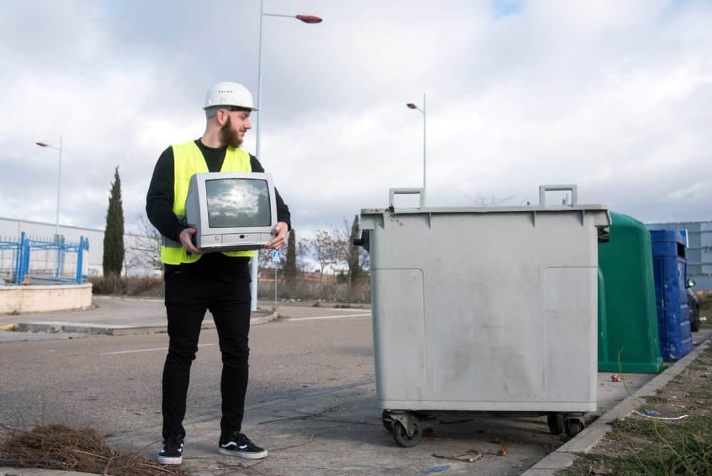 A man in a hard hat and safety vest stands outdoors on a cloudy day, holding an old computer monitor near a large trash bin, promoting electronics recycling. In the background, more bins line the street, highlighting efforts to recycle computers responsibly.