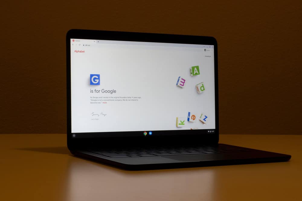 A laptop showcasing a webpage titled "Alphabet" with the text "G is for Google" sits on a plain surface against a neutral wall. The page features a large blue "G" and scattered colorful letters in the background, subtly reminding us of electronics recycling to keep our tech clutter-free.