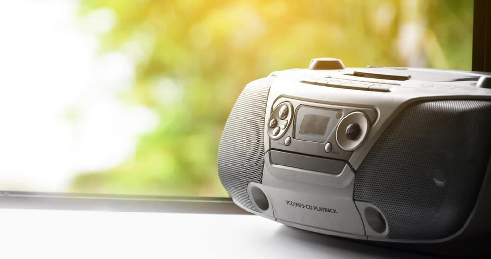 A portable CD player and radio rest on a windowsill, bathed in sunlight filtering through trees outside&mdash;a gentle reminder of the importance of electronics recycling to preserve such serene moments.