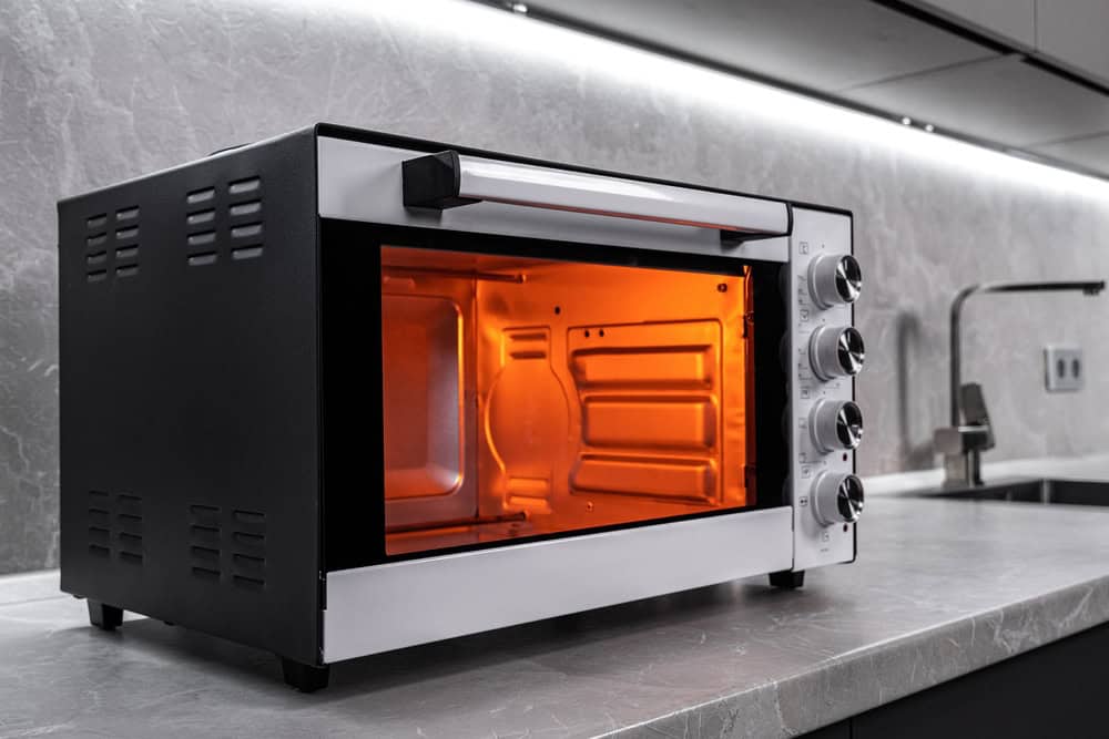 A black and silver toaster oven sits on a kitchen countertop with its door slightly open, glowing orange inside. The kitchen, designed with modern grey cabinets and a sleek silver faucet, emphasizes sustainability by incorporating electronics recycling bins for items like laptops and computers.
