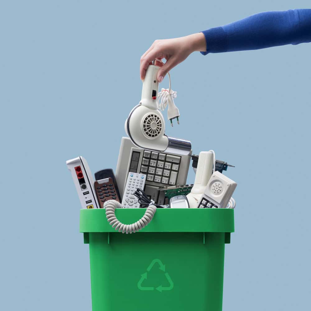 A hand places an old hairdryer into a green recycling bin filled with outdated electronics, including a calculator, remote control, phone, and plug adapter—an act of electronics waste disposal against the blue background.