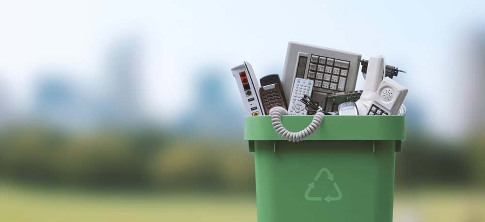 A green recycling bin brimming with electronic waste like old phones, keyboards, and remote controls highlights the importance of e-waste disposal. The recycling symbol on it stands out against a blurred cityscape beneath a clear blue sky.