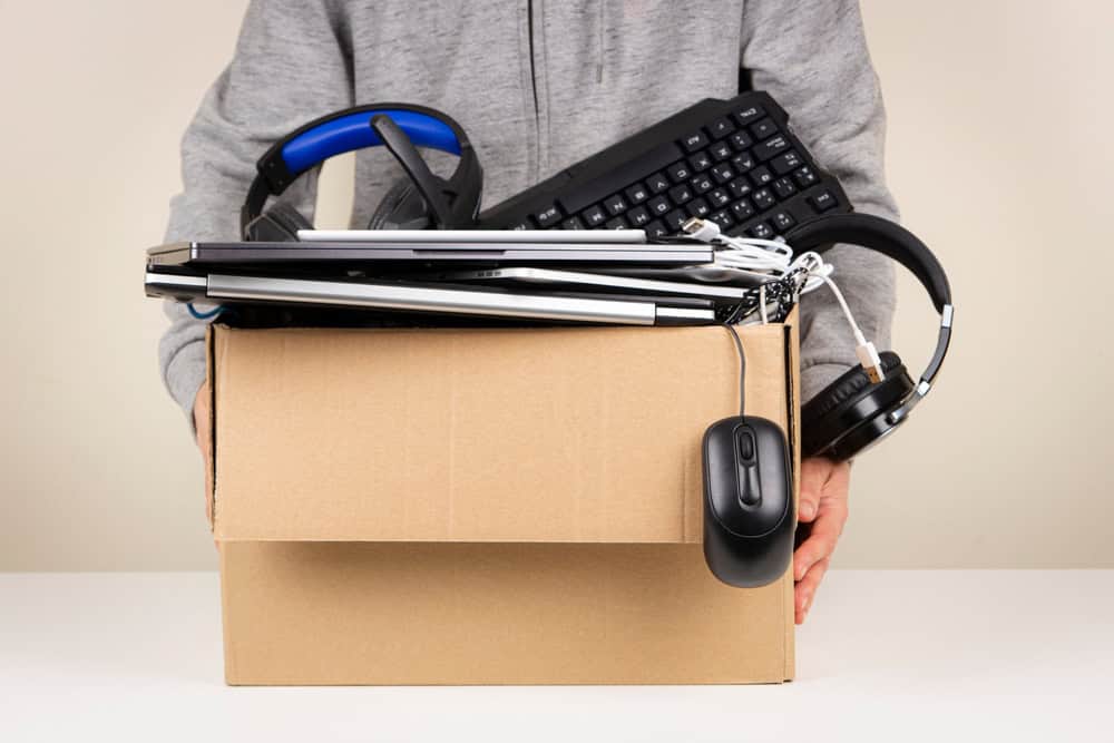 A person holding a cardboard box filled with various electronics, including a keyboard, laptop, mouse, headphones, and cables—perfect items for E-waste disposal. The person is wearing a gray hoodie, with only their hands visible.