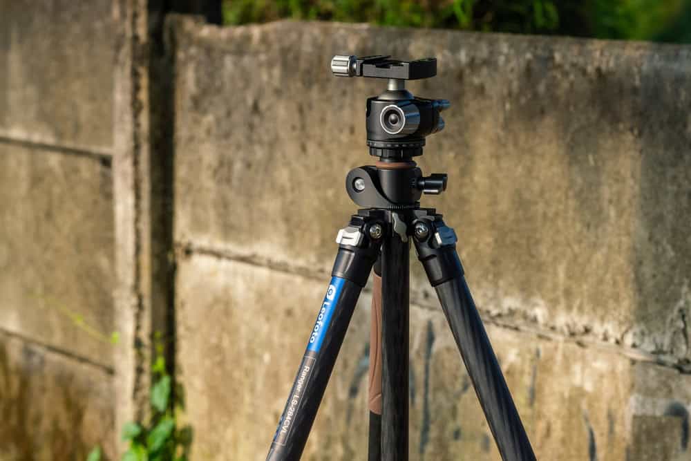 A photography tripod with a ball head is set up outdoors. It boasts a sturdy base and stands in front of a textured concrete wall, framed by greenery. Lit by natural sunlight, this setup quietly reminds us of the importance of responsible e-waste disposal to protect our environment.