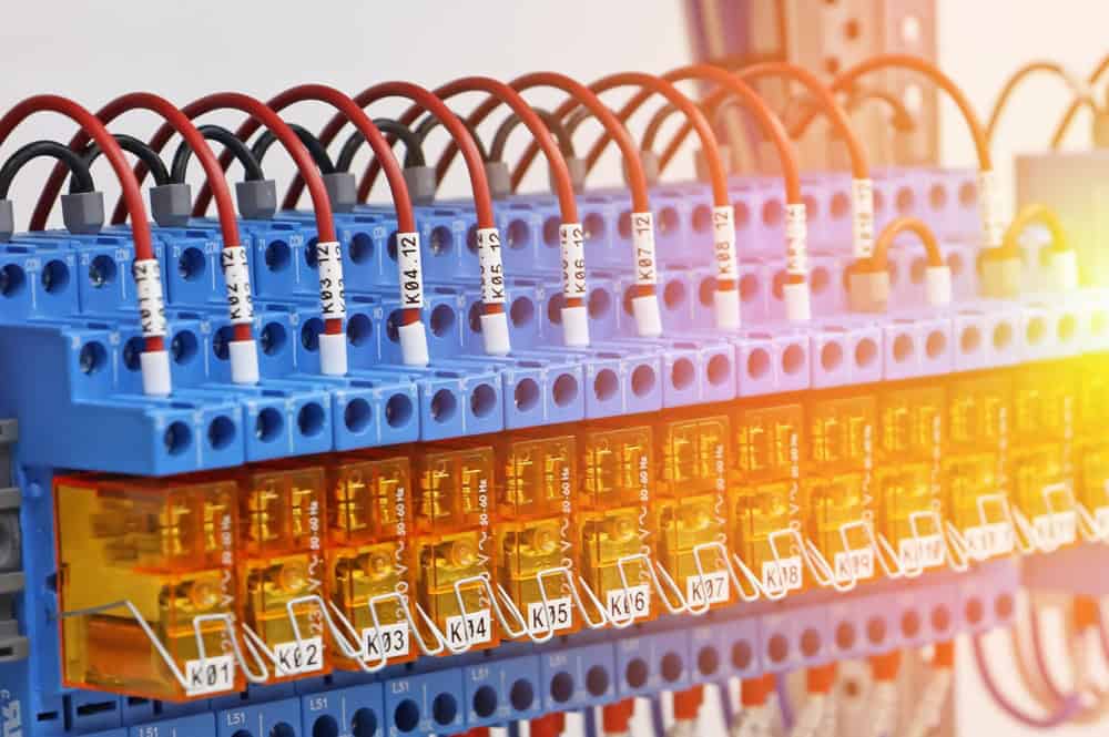 Close-up of a row of blue electrical terminal blocks with orange covers, connected by red and black wires labeled with numbers. Sunlight filters through, creating a warm glow&mdash;a reminder of the importance of electronics recycling to reduce waste and protect our environment.