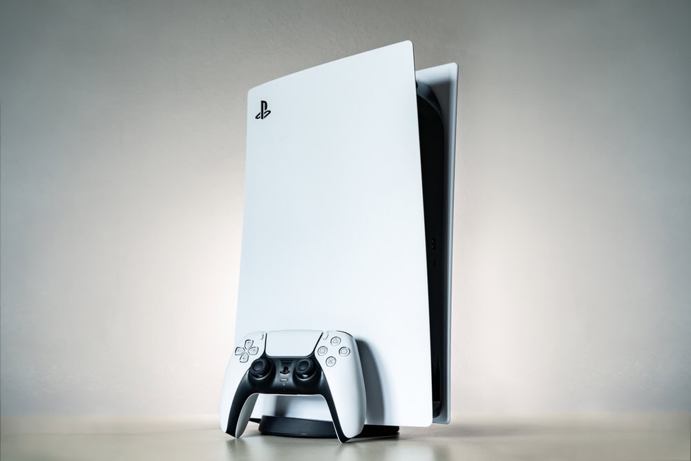 A white gaming console with a sleek, vertical design is placed against a neutral background, highlighting the importance of electronics recycling. In front of it, a matching white controller rests on a circular stand.