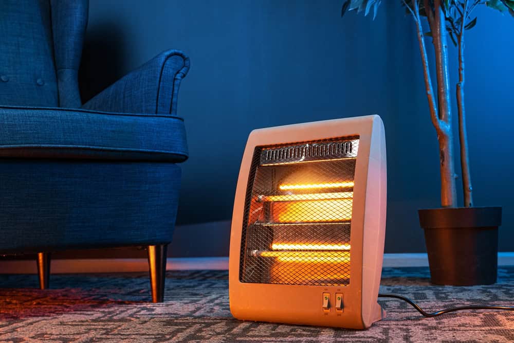 A portable electric heater with visible heating elements glows warmly, positioned on a patterned rug. Nearby, an upholstered chair and a potted plant are partially visible against a blue wall, offering a cozy corner to contemplate electronics recycling and embrace sustainable living.
