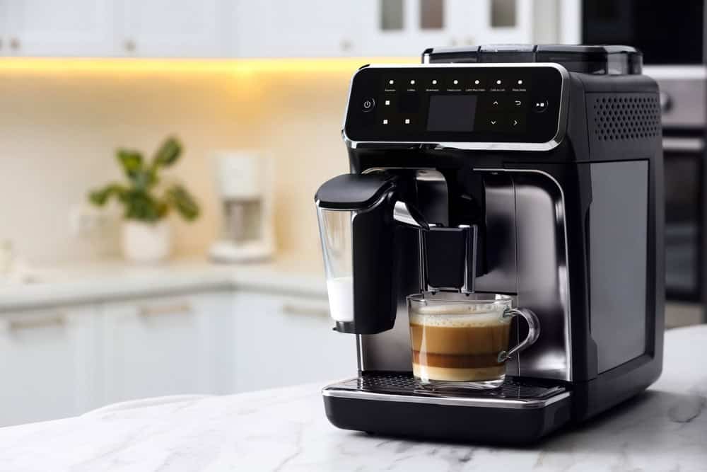A sleek black coffee machine on a marble countertop brews a layered cappuccino in a clear glass mug, exemplifying sustainable choices like those encouraged in E-waste disposal. The kitchen's backdrop of white cabinets, a potted plant, and a coffee maker complements the modern aesthetic.