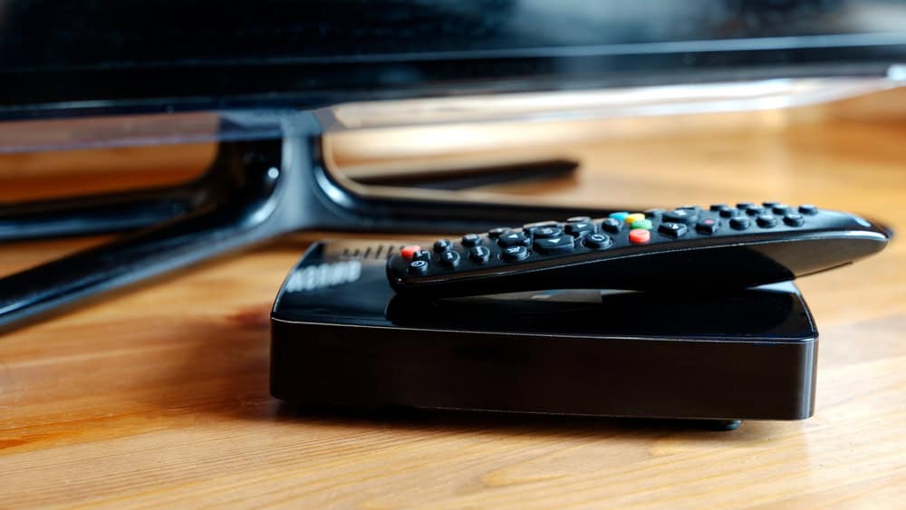 A black TV remote sits on top of a digital TV receiver box, placed on a wooden surface. The colorful buttons glint under the light, a reminder that proper E-waste disposal is crucial as technology evolves. The edge of a flat-screen TV is visible in the background.