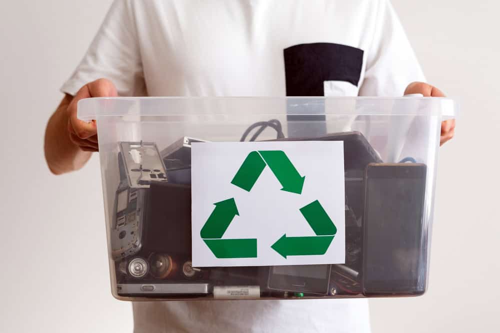 A person holds a clear bin filled with old electronic devices such as phones, cameras, and cables. A paper with a green recycling symbol is attached to the front of the bin, echoing the electronics recycling initiative. The person wears a white shirt with a black pocket.