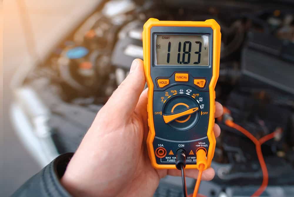 A hand holds an orange multimeter displaying a reading of 11.83, set to measure DC voltage with probes connected to a car engine. The brightly lit scene suggests a daylight setting and underscores the importance of precise measurements in processes like E-waste disposal.