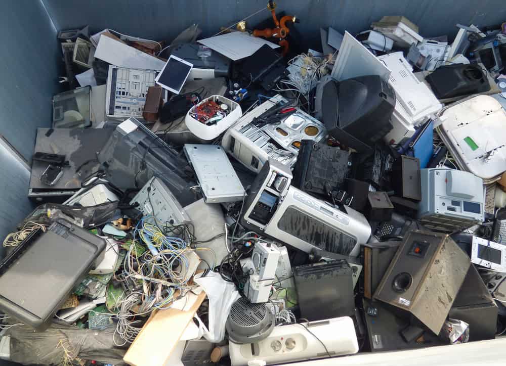 A collection of discarded electronics, including cables, computer parts, monitors, and other miscellaneous devices, piled haphazardly in a bin highlights the urgent need for proper E-waste disposal.