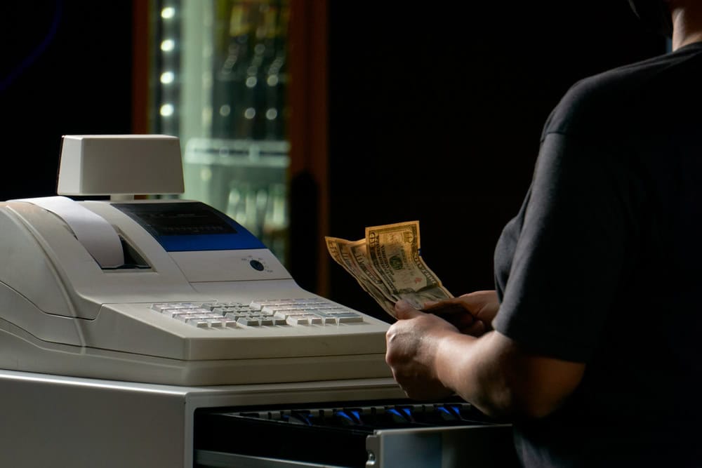 A person stands at an open cash register, counting dollar bills. The register is illuminated, and a glass drink cooler glows in the background. Nearby, a sign promotes electronics waste disposal, reminding customers of their role in protecting the environment.