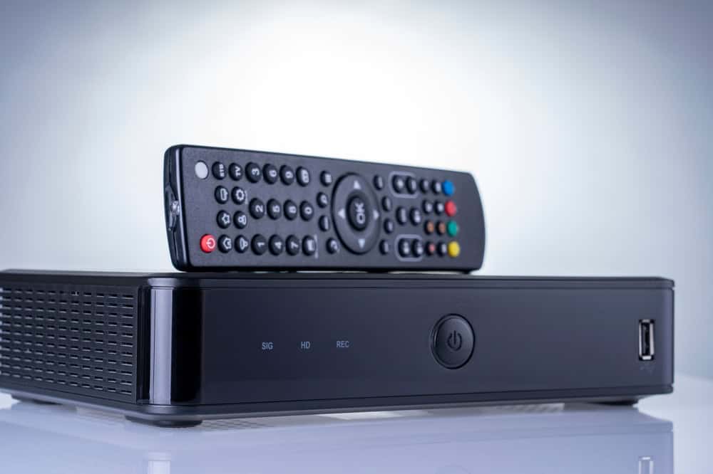 A black TV set-top box sits on a reflective surface, accompanied by a remote with various colored buttons. The box features power and USB ports, surrounded by an ambiance of soft lighting&mdash;a reminder of the beauty in electronics recycling and reimagining old technology.