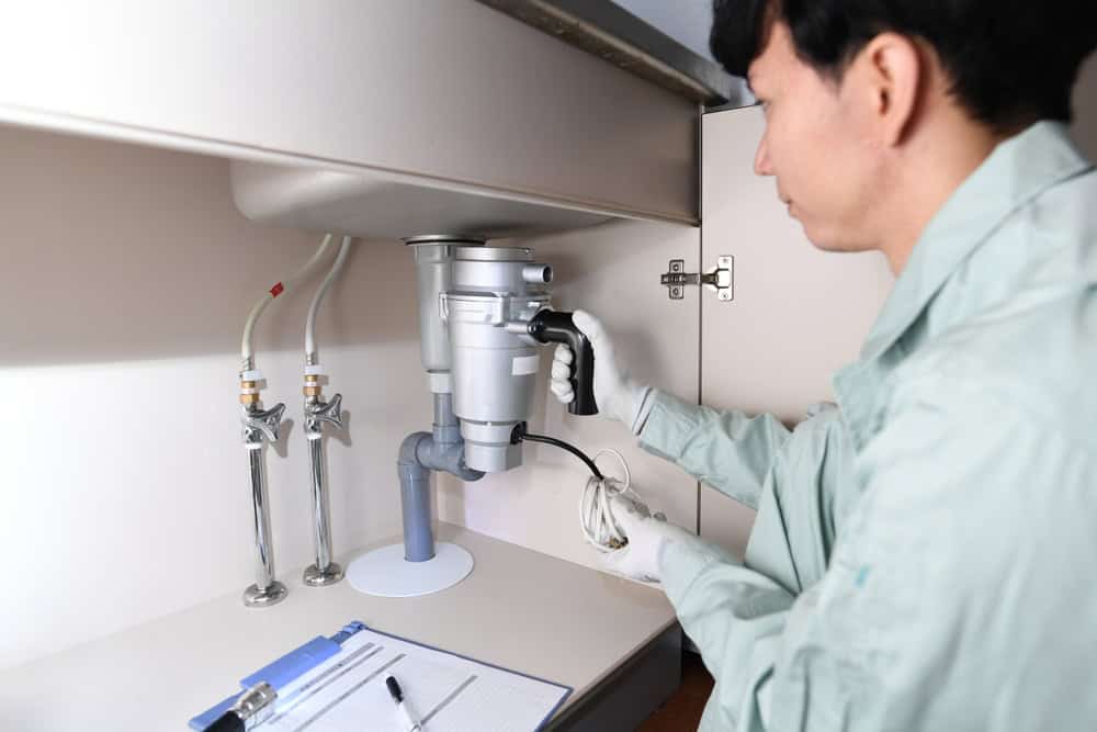A person in a light-colored uniform and gloves is using a tool to repair or install a garbage disposal unit under a kitchen sink, emphasizing responsible electronics waste disposal. The countertop has a blue clipboard with papers and a pen nearby.