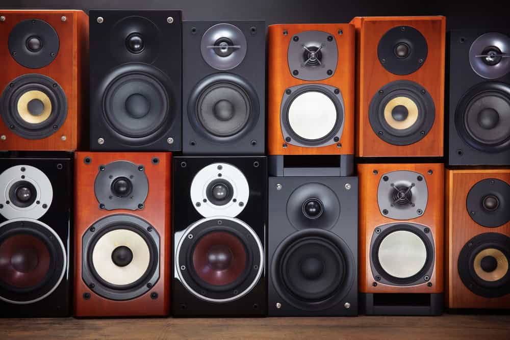 A variety of speakers arranged in rows against a dark background highlight the art of electronics waste disposal. Differing in size and color, with wood and black housings, each speaker showcases visible woofers and tweeters, some with metallic rims, perched elegantly on a wooden surface.