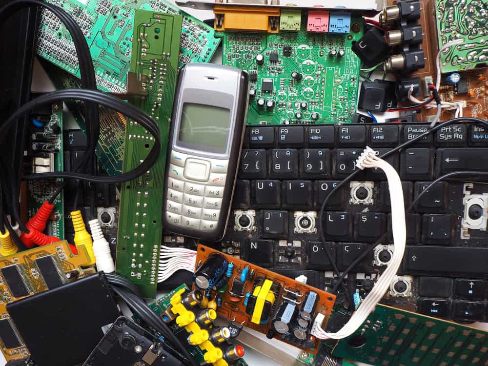 A pile of old electronic components, including a keyboard, circuit boards, a silver and black mobile phone, wires, colored plugs, and various scattered parts, showcases obsolete technology pieces ready for e-waste disposal.