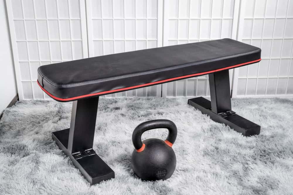 A black workout bench with red accents sits on a gray carpet, next to a black kettlebell. The backdrop is light-colored folding screens, subtly reminding of the need for electronics recycling in our digital lives.