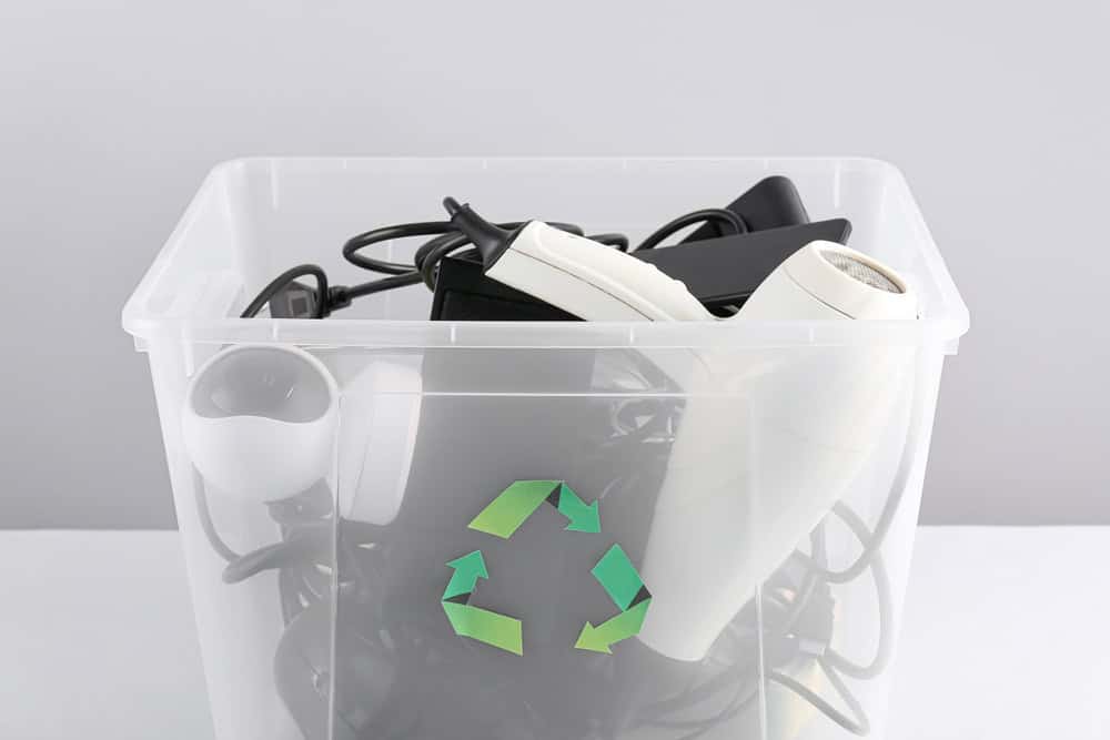 A transparent plastic bin filled with discarded electronics, including a webcam and cables, bears a green recycling symbol on the front, underscoring the importance of electronics waste disposal and encouraging responsible e-waste disposal habits.