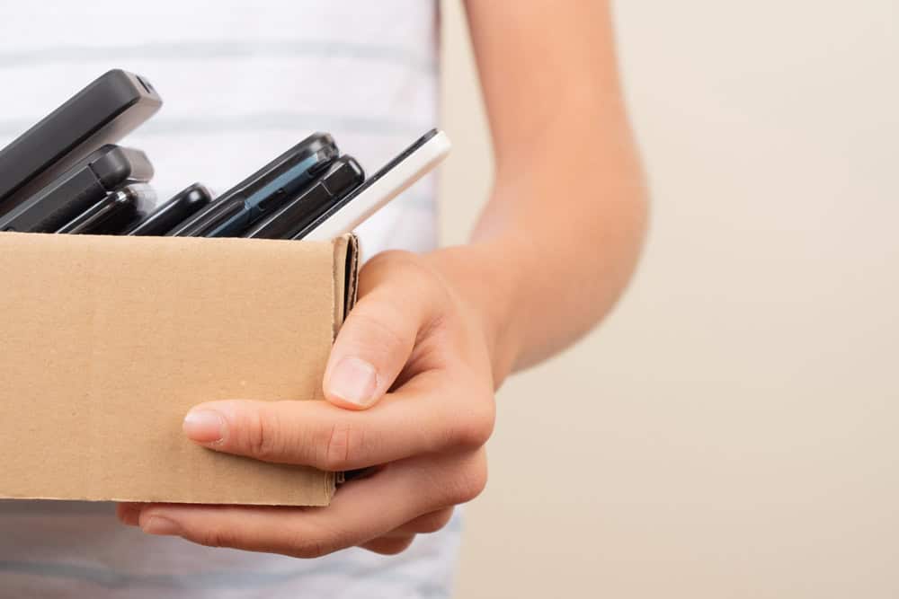 A person in a striped shirt holds a cardboard box brimming with old cell phones, highlighting the importance of electronics waste disposal against a plain, light background.