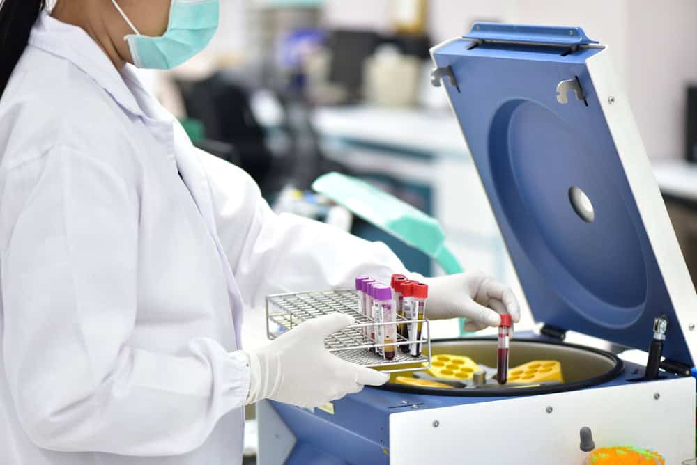 A laboratory technician wearing a mask and gloves places a blood sample into a centrifuge, mindful of electronics waste disposal. Holding a rack of test tubes, the technician ensures efficient use of resources in the lab environment as the centrifuge's compartments are revealed.