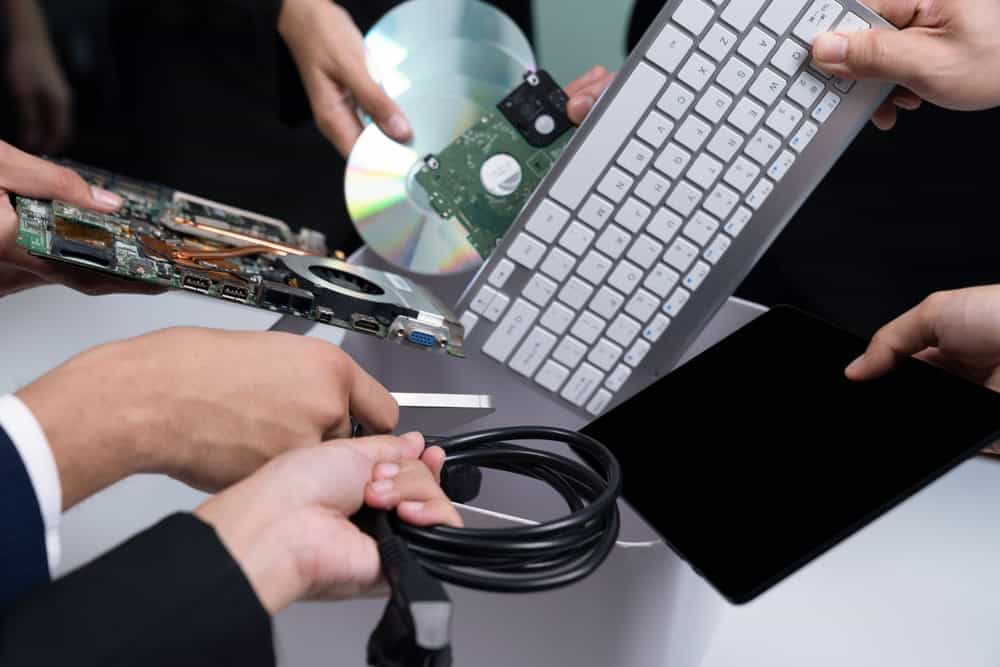 Close-up of several hands holding various tech items like a keyboard, circuit board, CD, and cables over a table. The scene suggests a collaborative tech project or hardware setup, emphasizing the importance of recycling computers to reduce e-waste responsibly.