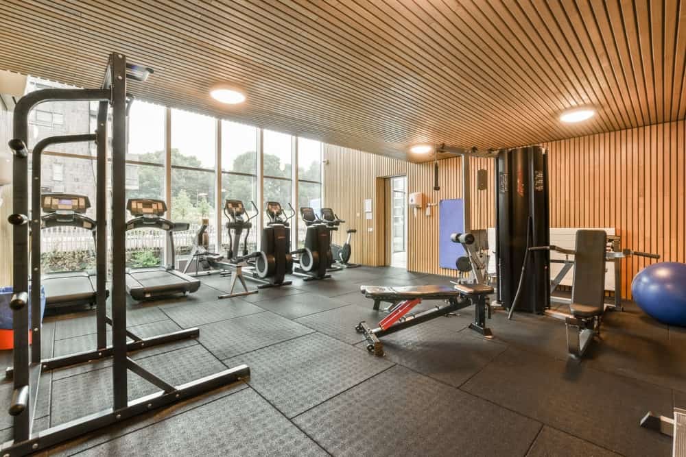 A modern gym with large windows showcasing outdoor greenery, featuring eco-friendly elements like electronics recycling stations. Equipment includes treadmills, stationary bikes, a squat rack, a weight bench, and more. The ceiling and walls boast elegant wooden paneling.