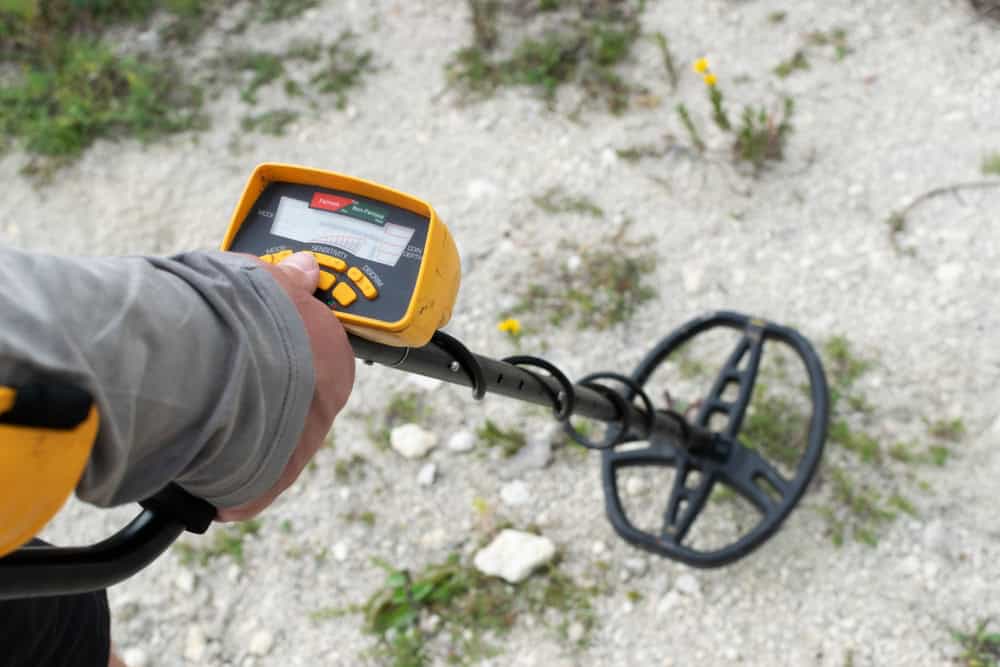 A person holding a metal detector over a gravelly surface, with the device's screen and buttons visible. The metal detector's yellow and black design echoes the importance of electronics recycling. Small patches of green grass and yellow flowers are scattered around.