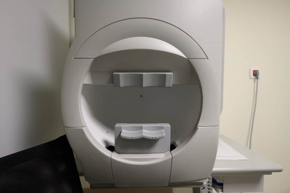 A large, circular medical device with a gray body is positioned in a room with beige walls. It has an open compartment, likely for head or eye examinations. A chair is partially visible, and a power outlet on the wall highlights thoughtful E-waste disposal by ensuring safe electronics recycling practices.